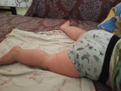 An indoor, close-up shot captures the lower body of a baby or toddler resting on a bed or sofa in Teykovo, Russia. The child is lying on their side, with their chubby legs and part of their torso visible. They are wearing light-colored shorts or underwear patterned with dark silhouettes of palm trees, dinosaurs, and surfboards, along with some numbers, including "433." A black waistband is visible, and a glimpse of a green or camouflage patterned garment can be seen on the right, likely part of the child's top.

The child is resting on a light-colored sheet or blanket with subtle, faded patterns. Beneath this, a darker brown patterned sheet or bedspread with intricate light-colored floral or paisley designs covers the surface of the bed or sofa. In the background, a blue and yellow patterned pillow is partially visible, alongside a brown pillow matching the bedspread's pattern. The scene depicts a peaceful moment of rest for the child. The time of day and weather are not discernible from the image.