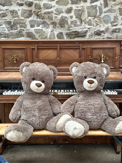 Two large, fluffy, brown teddy bears are seated side-by-side on a wooden bench in front of an upright piano. The piano has dark wood, a visible keyboard with black and white keys, and ornate decorative elements on its front. Behind the piano, a rough stone wall is visible. The lighting suggests an indoor setting, possibly a storage area or an artist's studio in Gonneville-la-Mallet, France. The teddy bears appear to be inanimate objects, posed for a photograph.