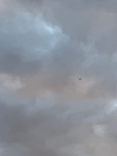 The image captures a dramatic sky filled with dense, grey clouds, suggesting overcast or stormy weather. A small, dark silhouette of an airplane is visible in the upper center of the frame, appearing to be in flight against the tumultuous backdrop. The airplane is small relative to the vastness of the sky, emphasizing its distance and isolation within the atmospheric conditions. The clouds are varied in texture and light, with some areas appearing brighter due to light filtering through, while others are deep and shadowed. The overall mood conveyed by the image is somewhat somber and awe-inspiring, highlighting the power of nature and the solitary presence of human technology within it. There is no visible text in the image. The location context provided is Drozhzhino, Russia.