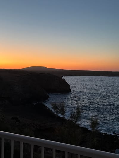 The image depicts a coastal sunset in es Mercadal, Spain. The scene is dominated by the ocean on the right and a dark, rocky shoreline on the left. The sky transitions from a light blue at the top to a vibrant orange and yellow near the horizon, indicating the sun is setting. A silhouetted mountain range is visible in the distance against the colorful sky. The water is choppy, with visible waves breaking along the shore. In the foreground, a white railing or balcony overlooks the scene, partially obscuring the lower portion of the rocky coast and some sparse vegetation. There are no people or discernible man-made structures in the natural landscape.