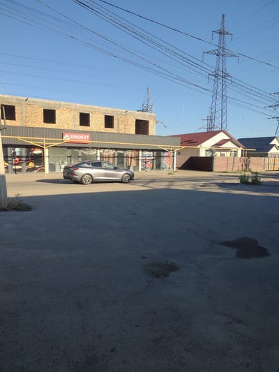 The image captures a street scene in the city of Zhaŋa Quat, Kazakhstan, under a clear blue sky. In the foreground, a grey sedan is parked on a paved surface. To the left, a building under construction displays exposed brickwork on its upper level, with dark, empty window frames. The ground floor of this building features storefronts with large windows, one of which displays a sign that reads "ERKIN ET". Adjacent to this building, a residential house with a red tiled roof and light-colored walls is visible. Further to the right, another house with a dark roof is partially seen. Overhead, power lines are strung between tall metal pylons, indicating the presence of significant electrical infrastructure. The overall impression is one of a developing urban or semi-urban area.