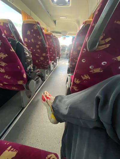 The image captures an interior view of a bus or train, oriented on its side (rotated clockwise). The scene shows multiple rows of passenger seats upholstered in a distinctive burgundy fabric with a repetitive pattern of light beige or gold abstract shapes and white oval-like elements. Each seat features grey armrests, and some have small grey tray tables or cup holders on the back.

In the foreground, a person's legs and feet are visible, indicating they are seated. The person wears grey trousers or track pants and colorful sneakers with prominent red, white, yellow, and gold accents. Their foot is slightly elevated or resting on the floor of the vehicle's aisle.

The floor of the vehicle is a light grey color with a speckled texture. Bright natural light streams in from windows along the left side of the vehicle (when upright), creating some glare. Overhead, the ceiling and internal structures are light-colored, possibly white or beige. Further down the aisle towards the front of the vehicle, more seats are visible, along with parts of the driver's area or cockpit through the front windshield, showing some dashboard elements and controls. A dark bag or backpack is tucked between two seats in the middle distance.

The lighting suggests it is daytime. No discernible text is visible within the frame. The location context provided is Duhovets, Bulgaria.