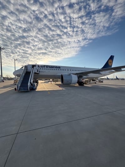 The image shows a Lufthansa Airbus A320neo aircraft on the tarmac at Munich Airport (MUC) in Oberding, Germany. The plane is connected to a set of mobile stairs, indicating passenger boarding or deboarding. The Lufthansa logo is visible on the plane's tail and fuselage. The sky is partly cloudy with a bright sun. The ground is concrete.