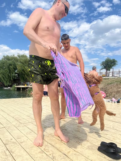 In Novomykolaivka, Ukraine, on a sunny day with scattered clouds, two men and a dog are on a wooden deck near a body of water. The man in the foreground, wearing black swim trunks with a repeating yellow Playboy bunny logo, is holding a purple and blue striped towel. He has a relaxed expression and is wearing sunglasses. To his right, a younger man is partially visible, also holding the towel. Behind them, a brown poodle-mix dog is lying down on the deck, with a pink and purple collar. In the background, there is a tree and a rustic structure near the water. Black flip-flops are placed on the deck to the right of the dog. The scene suggests a casual day at the beach or a lake.