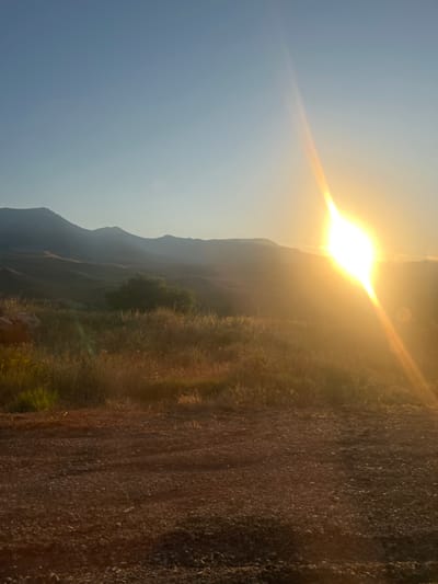 The image depicts a sunset over a rural landscape. The sun, a bright, glowing orb, is positioned in the upper right quadrant of the frame, casting a warm, golden light across the scene. A prominent lens flare emanates from the sun, stretching diagonally across the sky and a portion of the hills. In the middle ground, rolling hills are visible, their contours softened by the hazy light and distance. A solitary, dark green tree stands on one of the hillsides. The foreground is a rough, gravelly ground, with patches of dry grass and some greenery. The sky above is a clear, pale blue, gradually transitioning to a warmer hue near the horizon. The overall impression is one of a peaceful, natural setting bathed in the dramatic light of sunset. The location is the outskirts of Arpi, Armenia, characterized by its mountainous terrain and semi-arid vegetation.