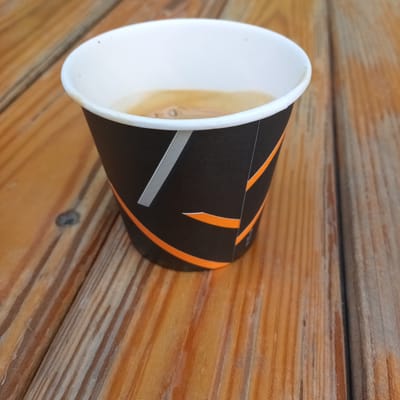 A close-up, high-angle shot shows a black and orange paper coffee cup on a wooden surface. The cup is partially filled with a light brown liquid, likely coffee. The wooden surface consists of planks with visible grain and some knots. The scene is set outdoors, possibly on a table or bench. The lighting appears to be natural daylight. There are no people or animals visible in the image. The overall impression is one of a simple, everyday scene.