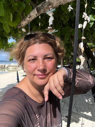 A woman with short, curly brown hair and freckles on her face is taking a selfie on a beach in Felidhoo, Maldives. She is wearing a sheer brown top with a floral print and sunglasses pushed up on her head. Her left hand is resting on her chin, and she is smiling. Behind her, there is a tree decorated with seashells, a black rope, and the ocean. The sand on the beach is light and appears to have footprints. The water is calm and clear, with some rock formations visible. The sky is blue with some clouds.