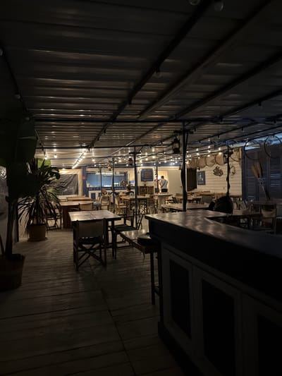The image depicts the exterior of a restaurant in Torreilles, France, at night. The scene is illuminated by string lights and lanterns hanging from the corrugated metal roof. The restaurant features wooden tables and chairs arranged on a wooden deck. In the foreground, a bar area is visible with a dark countertop and paneled base. Towards the back, there are people seated at tables, and a man in a white shirt and black pants is standing near a doorway. The restaurant appears to have a nautical or beach theme, with some decorative elements suggesting this, such as oars mounted on the wall.