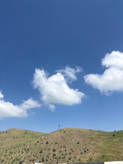 The image shows a wide view of a hilly landscape under a bright blue sky with scattered white clouds. The terrain is covered with sparse green grass, shrubs, and rocky patches. On the crest of one of the hills, a single, tall electrical pylon is visible. The scene suggests an outdoor environment in a rural or semi-rural area.