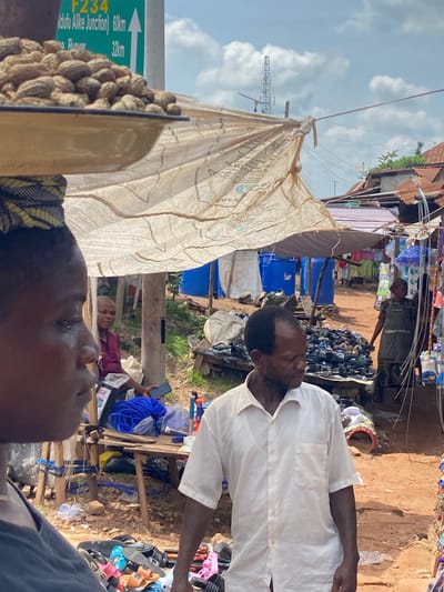 In Amaseri, Nigeria, a busy street market unfolds. In the foreground, a person is viewed from the side, their face obscured by the camera's perspective. They appear to be wearing a dark grey shirt. Behind them, a man in a light-colored, short-sleeved collared shirt stands facing right, his gaze directed downward. To the left, a vendor wearing a patterned headwrap balances a metal basin filled with peanuts on their head. A second woman, also wearing a headwrap and a maroon garment, is seated in the middle ground, holding a mobile phone. The market is characterized by makeshift stalls covered with tarps, displaying various goods including footwear, blue plastic barrels, and other merchandise. The ground is dusty and uneven, with scattered debris. In the background, a communication tower rises above the rooftops, and a green road sign indicates distances to Junction and Flower.