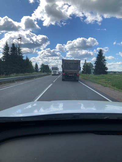 The image depicts a view from inside a vehicle traveling on a highway in the city of Dyatlitsy, Russia. The highway has two lanes in the direction of travel, separated by a white line. A guardrail runs along the left side of the road, and a grassy shoulder with trees borders the right. In the foreground, the white hood and windshield of the vehicle are visible.
Several trucks are present on the highway. The closest truck, in the right lane, is a dark-colored dump truck seen from the rear, with its license plate partially visible. Further ahead, in the left lane, a white semi-trailer truck is driving. The sky above is a bright blue, populated with fluffy white clouds.
The environment is rural, with dense green trees lining the road and fields of green grass and scattered houses visible in the distance on the right side. A tall communication tower with antenna elements is prominent in the left background, situated among the trees. The weather appears to be sunny with scattered clouds, suggesting a pleasant day.