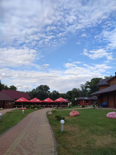 The image depicts an outdoor seating area of a restaurant or resort. A paved walkway curves through a grassy area, leading towards several wooden buildings with red roofs. Lined up in front of one of the buildings are numerous red umbrellas, under which tables and chairs are set up for dining. The umbrellas bear the "Coca-Cola" logo.
In the grassy area, there are decorative spherical objects and modern lampposts. Two large pink beanbag chairs are visible on the lawn, suggesting a relaxed atmosphere. The background is filled with lush green trees and foliage.
The sky is a vibrant blue with scattered white clouds, indicating pleasant weather, likely daytime. The architecture of the buildings, with their wooden log construction and dark roofs, suggests a rustic or traditional style. There is a small building with "KACA" written on it.
No specific activity or event is clearly depicted, but the setup implies a dining or leisure setting. The overall mood is tranquil and inviting. The location is identified as Shcherbani, Ukraine.