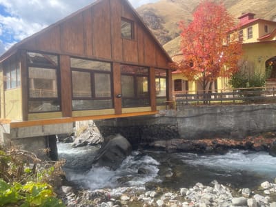 The image depicts a mountainous autumn scene in Juta, Georgia. In the foreground, a rapidly flowing river with visible white rapids and rock formations occupies the lower half of the frame, flowing from right to left under a cantilevered building.

To the left, a two-story structure with a brown wooden gabled roof and vertical wooden siding on the upper story extends into the frame. The lower story features pale yellow stucco walls and large, dark-framed windows forming a glass-enclosed space, which is supported by an orange structural beam and concrete piers extending into the river.

To the right, a second building with pale yellow stucco walls and a red tile roof is partially visible, featuring arched openings. Between these two structures, a deciduous tree with vibrant red-orange autumn foliage stands prominently. A low wooden fence runs along the top of a concrete retaining wall that borders the riverbank, separating it from the yellow-walled building and the red-leafed tree.

The background consists of steep, treeless, light brown mountains under a partly cloudy blue sky. No individuals or animals are visible in the scene.