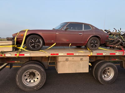 A maroon Datsun 280Z is secured to a flatbed trailer with yellow straps. A toolbox sits ahead of the car on the trailer, and a pile of bicycles is behind it. The trailer is part of a semi-truck, with a silver toolbox mounted between the sets of wheels. The wheels are large and show signs of wear. The sky is visible in the background.