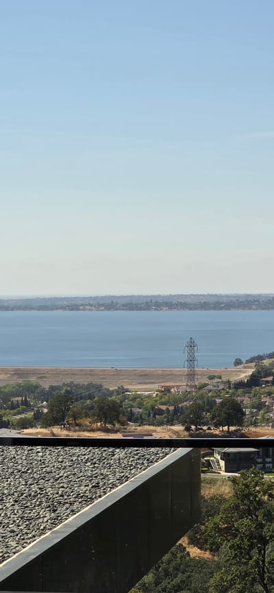 The image shows a scenic view of Folsom Lake in El Dorado Hills, California, United States. In the foreground, a modern architectural element with a pebble-covered surface and dark metal trim is visible. Beyond this structure, the landscape slopes down towards the lake. The midground features a wide expanse of the blue lake, bordered by a long, tan-colored dam or levee. A prominent electrical transmission tower stands near the dam, with a few dark-colored vehicles visible on the road atop the dam.

The far background displays a hazy outline of a city or town along the shoreline, with numerous buildings and trees. The sky is a clear, pale blue, indicating a sunny day with good visibility. The overall scene captures a picturesque blend of natural beauty and human infrastructure.