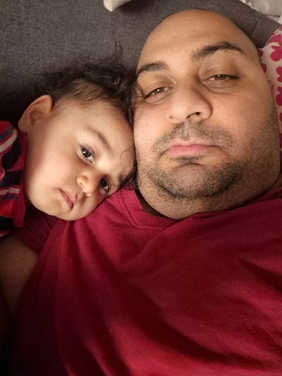 An adult male and a young child are depicted in a close-up indoor photographic capture, located in Melle, Belgium. The adult male, positioned on the right, is bald with dark stubble on his face, wearing a plain dark red crew-neck shirt, and directly faces the camera with a neutral, heavy-lidded expression. A young child, with dark, curly hair, rests their head against the adult male's left shoulder and chest area, looking towards the camera with a slightly upward and leftward gaze. The child wears a striped garment, showing red, white, and dark blue patterns on the visible left arm. The background behind the child's head is a dark gray fabric surface. To the right of the adult male's head, a white fabric with a pink patterned design is visible.