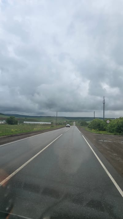 This image depicts a rural two-lane asphalt road in Кама-Исмагилово, Russia, under an overcast sky with heavy grey clouds. The road surface appears damp, suggesting recent precipitation, and is marked by solid white lines on the edges and a broken white line dividing the two lanes in the near foreground. A dark-colored sedan with its headlights illuminated is visible in the distance, traveling towards the viewer on the left lane.

To the left of the road, green fields with scattered yellow vegetation extend to a cluster of small buildings, including a long, low-slung white structure resembling an agricultural building, nestled against a backdrop of rolling green hills. Multiple wooden utility poles with overhead lines run parallel to the road.

To the right of the road, a dirt shoulder with a visible puddle transitions into an area of dense green bushes and small trees. A tall, open-lattice metal tower, likely for telecommunications, stands prominently. Adjacent to the tower, a circular white sign with a red border, indicating a speed limit of "50", is affixed to a pole. No people are visible in the scene.