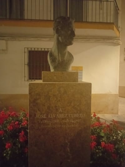 The image shows a bust of a man on a stone monument. The bust is made of dark stone and depicts the man in profile, looking to the right. The monument has text inscribed on it. The monument is located in a public space, likely a plaza or park, with red flowers in bloom around its base. In the background, there is a building with a balcony and a barred window. The overall lighting suggests it is nighttime or dusk. Based on the inscription on the monument, the location is Priego De Córdoba, Spain, and the monument is dedicated to A. José Álvarez Cubero, born in 1768 and died in 1827, described as "Homenaje de su pueblo" (Homage of his town).