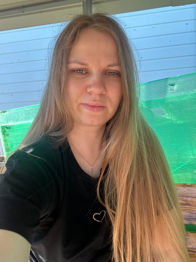 A close-up, waist-up selfie of a young woman with long, blonde hair and blue eyes is taken outdoors on a sunny day. She is wearing a black t-shirt with a small gold heart detail on the left side of her chest. A delicate gold necklace with a small pendant is visible around her neck. Her expression is neutral, with a slight smile. The background features a light blue, corrugated surface overhead and a bright green, corrugated plastic barrier to her right. The environment appears to be in the city of Lotoshino, Russia, possibly a rural or semi-rural setting given the rustic wooden elements visible behind the green barrier.