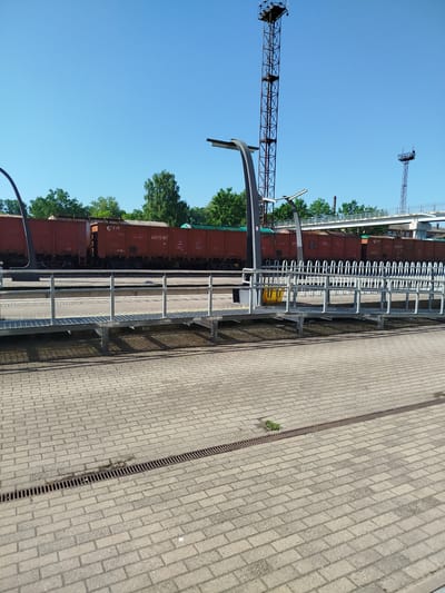 The image depicts a sunny day at a train station in Valga, Estonia. In the foreground, a paved area with a drainage grate runs across the frame. Beyond this, a metal walkway with railings leads towards a train track where several red freight cars are parked. In the background, a clear blue sky is visible above a line of trees. Two tall metal towers with lights, characteristic of a train station, stand against the sky, one on the left and a smaller one on the right. A pedestrian bridge is also visible in the distance. No people are present in the scene.