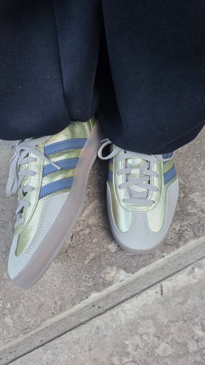 A close-up, eye-level shot focuses on a person's feet clad in gold and blue Adidas Sambas, standing on a textured concrete surface. The shoes are a metallic gold with pale blue stripes and grey laces. The person is wearing dark, baggy pants that obscure most of their body. The background shows more of the concrete surface, which appears to be outdoors, possibly a sidewalk or a step. The setting is the city of Рассудовец Снт, Russia.