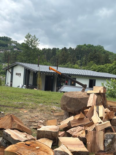 In Kapreshumi, Georgia, a scene unfolds with a modern building with the words "STONE RIVER" prominently displayed on its exterior. In front of the building, a large tree stump serves as a chopping block, with an axe firmly embedded in it, its orange handle contrasting with the weathered wood. A substantial pile of freshly cut logs surrounds the stump, indicating recent activity. The environment is lush and green, with trees and vegetation covering the rolling hills in the background. The sky is overcast, suggesting a possibility of rain.
