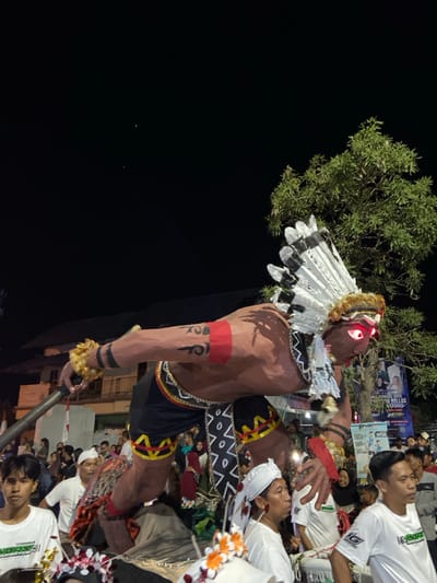 A large-scale cultural float dominates the scene at night in Kedungbanteng, Indonesia. The float depicts a muscular, dark-skinned male figure in a dynamic, crouching pose, extending its right arm forward. It wears a prominent white and black feathered headdress, a dark sarong-like garment with yellow and red geometric patterns, and various armbands, including one with yellow fur/feathers. The figure's eyes glow red, and tribal-style markings are visible on its arms.

The float is positioned amidst a crowd of people. Several individuals are visible in the foreground and mid-ground, some wearing white shirts. One man in a white shirt and white headwrap stands directly in front of the float, looking left. To his left, a young man in a white t-shirt with green text looks towards the viewer. To the right, another man in a similar white t-shirt walks right. A dense crowd occupies the background.

The environment is an urban setting at night, illuminated by artificial light. A large leafy green tree is visible on the right, partially obscuring a building. On the left, a multi-story building and a flagpole displaying a red and white flag are present. A large banner or advertisement is visible in the background on the right.