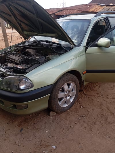 A light green Toyota Avensis station wagon, with its hood open, is parked on a dirt surface in Kontagora, Nigeria. The car's driver's side door is also open. The engine bay is visible, revealing the engine, battery, and other components. The car is positioned on a road or area with loose soil and scattered debris, with a wooden pole visible to the left and a building with a corrugated metal roof in the background. The tire on the front driver's side is visible, as is the front headlight and bumper. The scene appears to be outdoors, likely in a residential or semi-urban area given the building in the background.