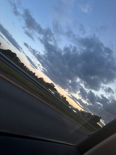 The image shows a view from a vehicle traveling on a highway at dusk. The sky is a gradient of blues and grays with scattered clouds. The sun is low on the horizon, casting a warm glow through the clouds. On the left side of the image, trees line the highway. The highway itself is dark asphalt with a yellow line marking the lane. In the distance, lights from buildings or vehicles can be seen. The image is tilted, suggesting it was taken while the vehicle was in motion. There are no people or specific identifiable landmarks that would indicate a location in Missingmyr, Norway.