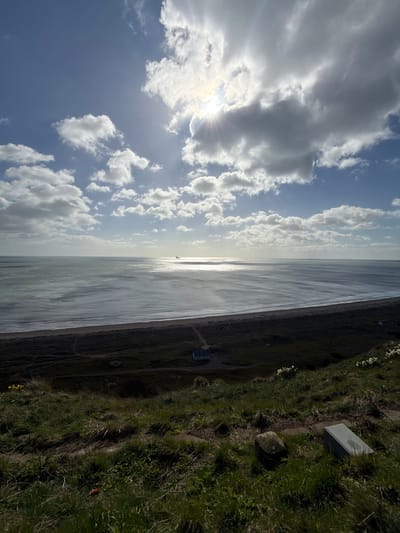 The image shows a view from Montrose, United Kingdom, depicting a shoreline scene. The horizon line divides the image in half, with a cloudy sky above and the North Sea below. The sun is visible through the clouds, creating a bright glare. The sea is calm, with a strip of beach separating it from a grassy coastal area. The grassy area rises slightly, leading to a flat area where a small building and stone structure stand. The image is taken from a high vantage point, and the angle is rotated 90 degrees.