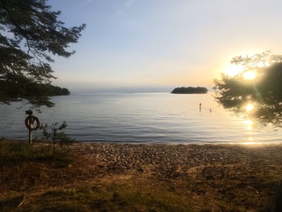 This media file depicts a tranquil sunset scene at a beach, likely located in Motala, Sweden. The setting is a serene body of water, possibly a lake or the sea, with calm ripples reflecting the warm hues of the setting sun. In the foreground, a sandy beach stretches out, with sparse vegetation and pine trees framing the scene. A life preserver is mounted on a post near the water's edge, suggesting a public swimming area.
Several figures are visible in the water, appearing to be bathing or wading, enjoying the last moments of daylight. One person stands further out in the water, while two others are closer to the shore. The sun, a bright orb of light, is low on the horizon, casting a golden glow and creating a shimmering path across the water. The sky transitions from a pale blue at the top to a soft yellow and orange near the horizon, indicating the late evening hour and a clear, calm weather.
The overall mood of the image is peaceful and idyllic. The warm, diffused light and the presence of people enjoying the water evoke a sense of relaxation and simple pleasure. The natural beauty of the landscape, combined with the serene activity, creates a picturesque moment frozen in time. There is no visible text in the image.