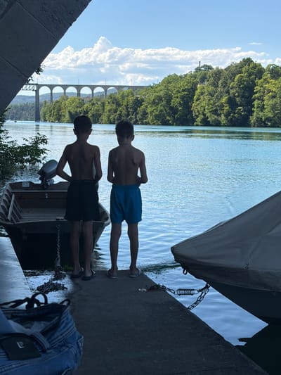Two young boys, silhouetted against a bright blue sky and a wide body of water, stand on a concrete dock. They are facing away from the camera, looking out at the water. The boy on the left wears dark shorts, and the boy on the right wears blue shorts. To their left, a small, empty boat with an outboard motor is tied to the dock. To their right, a covered boat is moored. In the background, a lush green forest lines the opposite bank of the river, and a large, arched stone viaduct spans the water in the distance. The scene is likely the Rhine River in Eglisau, Switzerland, a town known for its picturesque river and historic viaduct. The lighting suggests it is a sunny day.