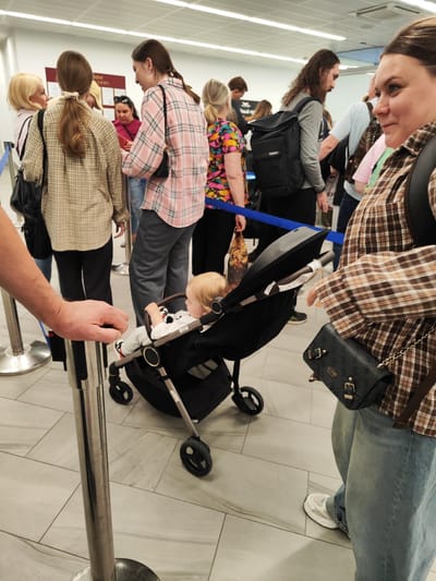 In Khrabrovo, Russia, at what appears to be an airport or similar travel hub, a group of people are standing in line. In the foreground, a person's hand is resting on a stanchion, and next to them, a baby is seated in a stroller. The baby is facing away from the camera, looking at the stroller's handle. Further back in the queue, individuals are dressed casually, some wearing plaid shirts, and one person is carrying a backpack. The environment features a tiled floor and a suspended ceiling with fluorescent lighting. A blue rope delineates the queue.