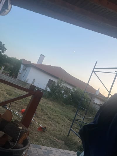 The image depicts a residential area in Sokolovo, Bulgaria, during the late afternoon or early evening. A white house with a red-tiled roof is visible in the background, featuring a prominent chimney. In the foreground, a wooden railing and a pile of chopped firewood are present. Two orange and silver grass trimmers are lying on the dry grass, suggesting recent or ongoing yard work. A metal scaffolding structure stands to the right of the house. The sky is clear with a faint crescent moon. The overall scene conveys a quiet, rural atmosphere.