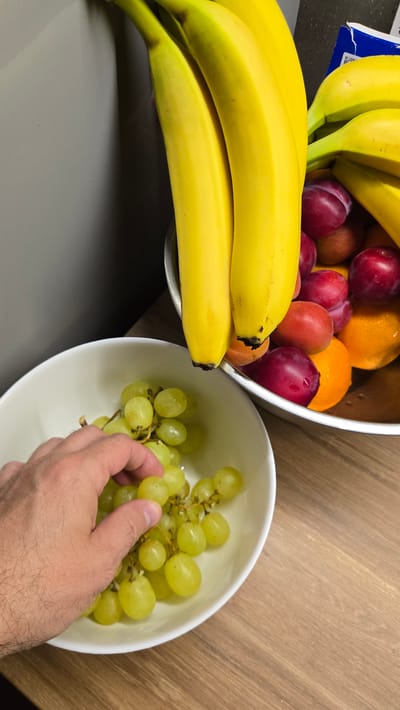 This image shows a close-up of a person's hand reaching into a white bowl filled with green grapes. The hand is picking a bunch of grapes. In the background, there is another bowl filled with bananas, plums, and oranges. The bowls are on a wooden countertop, and the background wall appears to be grey. This scene is a domestic setting, likely a kitchen, and there is no indication of the city of Lebedyan, Russia, or any specific forensic details.