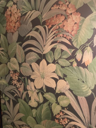 The image shows a close-up view of a wallpaper with a tropical floral and foliage pattern. The design features large, stylized leaves in various shades of green and beige, interspersed with clusters of small, coral-colored flowers and larger, pale yellow flowers. The background is a dark, solid color, likely charcoal or black, which makes the vibrant colors of the plants stand out. The wallpaper appears to be textured, with a slight raised effect on some of the floral elements. The lighting is soft and diffused, creating subtle shadows and highlights that give the pattern depth. The image does not depict any specific location in Gerstheim, France, as it is a close-up of a decorative surface.