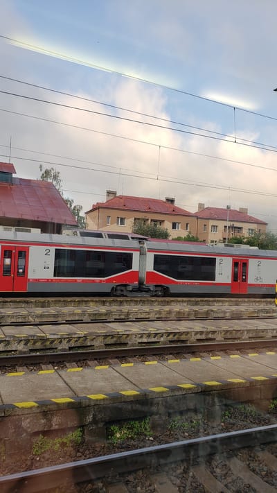The image shows a train at a train station in Margecany, Slovakia. The train is red and white and appears to be modern. The train station has multiple tracks and platforms, and there are buildings in the background. The sky is cloudy. There are no people visible in the image.