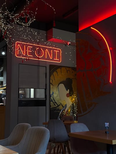 The media file shows an interior scene with a focus on a neon sign that reads "NEONI". The sign is illuminated with a bright orange-red light and is positioned vertically. Surrounding the sign are branches adorned with small white fairy lights, creating a festive and somewhat whimsical atmosphere. To the left of the neon sign, there is a large red triangular shape, likely a wall or architectural element, illuminated by a red light. In the background, a large mural or artwork is visible, depicting stylized figures in red and gold tones against a dark background.
Further to the right, there are glimpses of furniture, including what appear to be chairs upholstered in gray fabric. One chair, in particular, has a distinctive ribbed texture on its backrest. A wooden table or desk is also visible, with a small object attached to its underside, possibly a QR code or a small decorative item. The overall lighting suggests an indoor setting, possibly a restaurant, cafe, or entertainment venue, with the neon sign and fairy lights contributing to the ambiance. The perspective is angled, looking upwards and to the side. The image has a slightly grainy quality, suggesting it was taken with a smartphone camera in low light conditions.