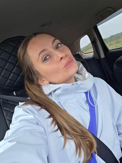 The image displays a woman from an overhead, slightly rotated perspective, likely a selfie taken inside a car. The woman has light skin, striking blue eyes, and long, light brown hair that flows over her shoulder. She is looking directly at the camera with a calm and neutral expression, her lips slightly parted. She wears a light-colored, possibly white or pale blue, hooded jacket with visible buttons and a zipper. A bright purple strap is visible across her chest, under a black seatbelt that diagonally crosses her torso, indicating she is secured in the vehicle.
She is seated in a black car seat featuring a quilted leather design. Through the car's side window and what appears to be a sunroof, a rural landscape unfolds. This external view shows wide, green or brown fields under an overcast, grey sky, suggesting a cloudy day. The setting appears to be a natural, open environment, consistent with the provided location context of Бабстово, Russia. No visible text is present in the image. The scene conveys a sense of a quiet journey or a brief stop during an overcast day.