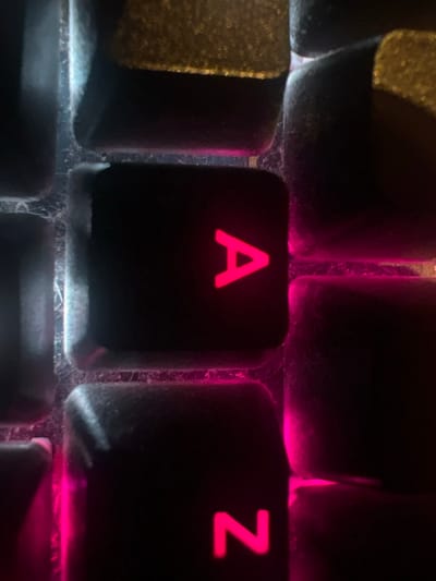 A close-up view shows illuminated keyboard keys. The "A" and "Z" keys are prominently featured, glowing red. The surrounding keys are dark, with some reflecting light. Dust and grime are visible between the keys, giving the keyboard a used appearance. The focus is sharpest on the "A" and "Z" keys, with the surrounding area appearing slightly blurred. Overall, the image highlights the contrast between the illuminated keys and the dirty keyboard surface.