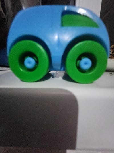A blue toy car with green wheels and window is positioned near a white wall. The wheels have a distinct blue center. A shadow of the toy is cast on the wall, creating a soft contrast. The lighting appears to be coming from the left side of the image, highlighting the curves of the car. The background is slightly blurred.