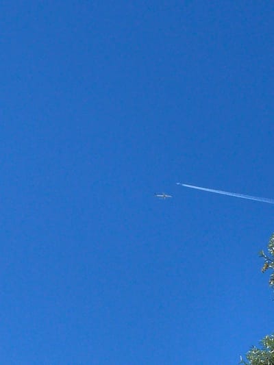 This media file captures a clear blue sky with a notable feature: a plane leaving a distinct contrail. The plane itself is small and white, appearing as a tiny speck against the vast expanse of the sky. The contrail stretches diagonally upwards and to the left, suggesting the plane's flight path. In the upper right corner, a portion of lush green foliage from a tree is visible, indicating an outdoor setting on a bright day. The leaves are detailed and appear healthy, with sunlight dappling through them. The overall scene is peaceful and expansive, evoking a sense of calm due to the clear sky and the distant, unobtrusive presence of the aircraft. The lighting suggests it is daytime, likely late morning or early afternoon, given the brightness and clear visibility. There is no visible text in the image. The location context provided is Lokeren, Belgium, which might imply a common scene for people in that area.