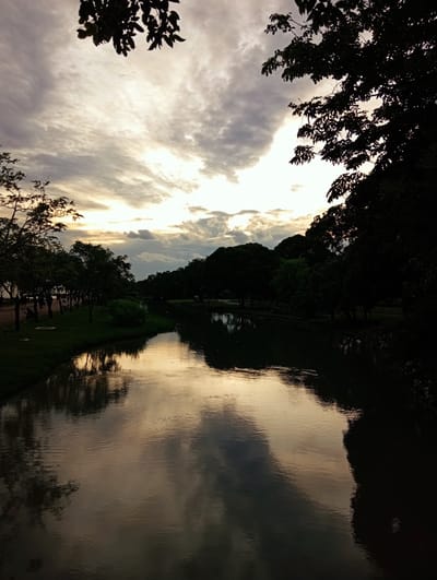The image depicts a serene canal scene in Bang Kaeo Subdistrict, Thailand, during dusk. The water's surface reflects the dramatic, cloud-filled sky, painted with hues of yellow and grey. Dark silhouettes of trees line both sides of the canal, with the foliage on the right casting a deep shadow across the water. On the left bank, a grassy area is dotted with trees and a few indistinct figures are visible walking or sitting, suggesting a park or recreational area. The overall atmosphere is tranquil, bathed in the soft, diffused light of the setting sun.