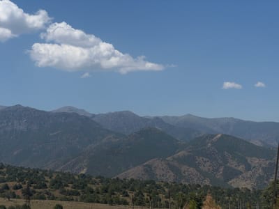 The image shows a vast mountainous landscape under a clear blue sky with scattered white clouds. The foreground is covered with low-lying vegetation and scattered trees. Further back, rolling hills rise, dotted with more trees and bushes, some of which appear to be scrubland. In the distance, several layers of progressively hazier mountains recede towards the horizon. The overall scene depicts a rural, natural environment with no signs of human habitation or activity. The location is identified as the city of Dardar, Uzbekistan.