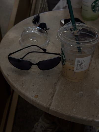 The image shows a close-up, rotated view of a Starbucks cup with an iced latte, a pair of sunglasses, and a small glass jar on a textured, speckled marble surface. The Starbucks cup is partially filled with an iced latte and has a green straw. A white sticker on the cup reads "Cafe To Go," "Item: 2 of 2," "DANIA," "GR ICED LATTE," "Time: 01:36:51 PM," and ">Starbucks< 4939." The sunglasses are black and lie next to the cup, with their lenses facing upwards. The glass jar is small and clear, positioned between the cup and the sunglasses. The marble surface appears to be part of a table. The lighting suggests it is daytime. The scene is indoors, on a table, possibly in a cafe or a home setting. There are no people visible in the image.