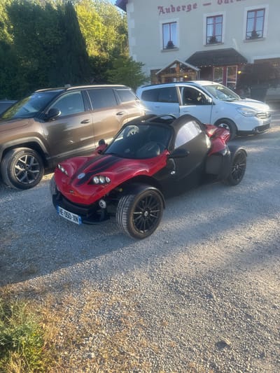 In Charchilla, France, a three-wheeled red and black sportscar is parked on a gravel lot in front of a building that appears to be an inn or restaurant, indicated by the signage "Auberge - Durande". Two other vehicles, a brown SUV and a white van, are also present. The sportscar has a black convertible top and prominent headlights. A license plate reading "HE-050-XN" is visible on the front of the sportscar. The environment suggests a sunny day with trees and vegetation in the background. There are no people visible in the image.