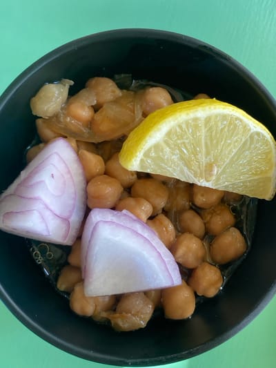 A close-up, top-down view captures a black, circular bowl containing a dish of cooked chickpeas, captured in Platys Gialos, Greece. The chickpeas, light brown and rounded, are partially submerged in a clear, oily liquid, intermingled with pieces of softened, translucent onion. Two prominent wedge-shaped slices of red onion, displaying white and purple concentric rings, are positioned in the lower-left section of the bowl. A bright yellow lemon wedge, cut lengthwise to reveal its pulpy interior, rests on the chickpeas in the upper-right. The bowl is situated on a solid, bright green surface, under even illumination that produces minimal shadows. No human presence or direct action is depicted.