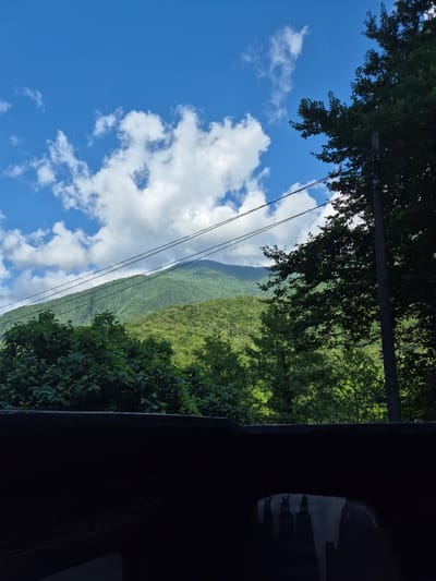 This media file depicts a lush, green mountainside under a bright blue sky dotted with fluffy white clouds. The perspective is from an elevated position, possibly looking out from a building or a vehicle, with dark, silhouetted structures framing the right side of the image. In the foreground, the dark mass suggests an interior space or an overhang, with a reflection visible on its surface that is abstract and indistinct.

Dominating the left side of the frame is a vibrant blue sky filled with cumulus clouds, some appearing bright and sunlit, others with softer edges. A power line, consisting of two dark cables, stretches diagonally across the sky and into the distance, partially obscuring the view of the mountains.

The mountains themselves are covered in dense, verdant vegetation, with layers of trees creating a textured and undulating landscape. The sunlight appears to be strong, casting a clear light on the scene and highlighting the rich green hues of the foliage.

The time of day appears to be daytime, with the bright sky and sunlight. The weather is clear and pleasant, indicated by the blue sky and clouds. The location cues, such as the power line and the densely vegetated mountainside, suggest a rural or natural setting, possibly in a mountainous region.

The overall impression is one of natural beauty and tranquility, with the dramatic landscape contrasted by the man-made element of the power line and the framing of the dark foreground. There is no visible text in the image.