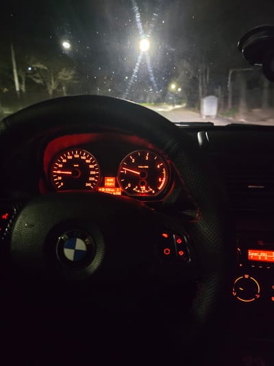 A night-time scene is depicted from the interior perspective of a BMW vehicle, positioned in Kumanovo, North Macedonia. The vehicle's dashboard is illuminated in red, displaying a speedometer indicating 40 km/h and a tachometer registering approximately 1500 RPM. A central digital display shows the time as 19:03, an outside temperature of -12.0°C, a remaining range of 530 km, and a trip meter reading of 294. The black, textured steering wheel, featuring red stitching on the right side, prominently displays the BMW emblem. Illuminated control buttons are visible on the left spoke of the steering wheel. To the right, a portion of the center console features an orange-illuminated radio display reading "Tuner FM1" and an illuminated climate control dial.

The view through the windshield reveals a dark, potentially wet, road ahead. Multiple external light sources, including streetlights, are visible, creating glare and reflections on the windshield, which appears smudged or wet. Sparse trees and faint outlines of structures or fences line the sides of the road in the distance, indicative of a semi-urban or suburban environment. A black suction-cup mount is affixed to the upper right corner of the windshield. No individuals are visible. The speedometer reading confirms the vehicle is in motion.