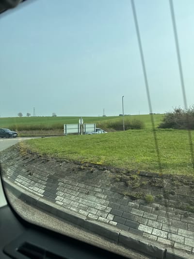 This image captures a wide-angle view from inside a vehicle at a roundabout on the outskirts of Thrapston, United Kingdom, likely within the A14/A45/A605 junction complex.
In the immediate foreground, a grey concrete retaining wall with a distinctive pattern of alternating light and dark rectangular blocks extends horizontally. Sparse grass and weeds grow along its top edge. A dark grey curb delineates the wall from a visible section of dark, damp-appearing asphalt road surface at the bottom of the frame. Vertical light streaks, consistent with reflections on a windshield, are visible in the upper right.
The midground features a large, green grassy island, characteristic of a roundabout's center. On the left side of the image, a dark-colored SUV, possibly black, is observed on the asphalt road, indicating movement from left to right around the roundabout. Positioned centrally on the grassy island are two white rectangular road signs mounted back-to-back on a single pole. Behind these signs and slightly to their right, a silver or light-colored sedan is partially visible, resting on a grassy incline with only its roof and upper body section protruding above the grass. A tall, dark grey lamppost stands vertically on the right side of the grassy island, adjacent to a dense mass of dark green shrubs or bushes.
The background reveals expansive green agricultural fields under a hazy, light blue sky. Multiple electricity transmission pylons are distributed across the distant landscape, with several clusters of deciduous trees visible along the horizon line. No individuals are present. The scene is illuminated by bright, diffuse daylight.